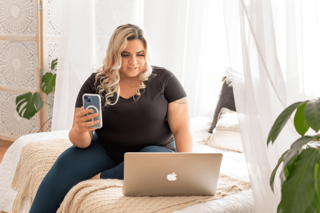 A woman doing work on both her cellphone and laptop while seated on her bed.